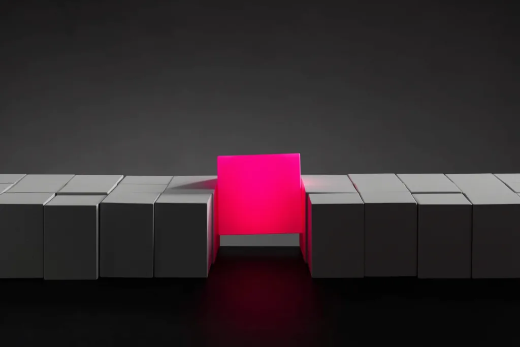 Identical grey cubes with one coral cube stepping forward on a dark surface.