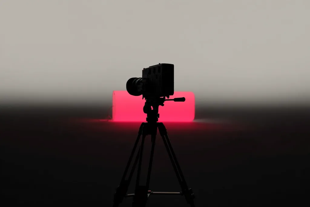 A cinema camera silhouetted with a coral light behind it on a dark set.