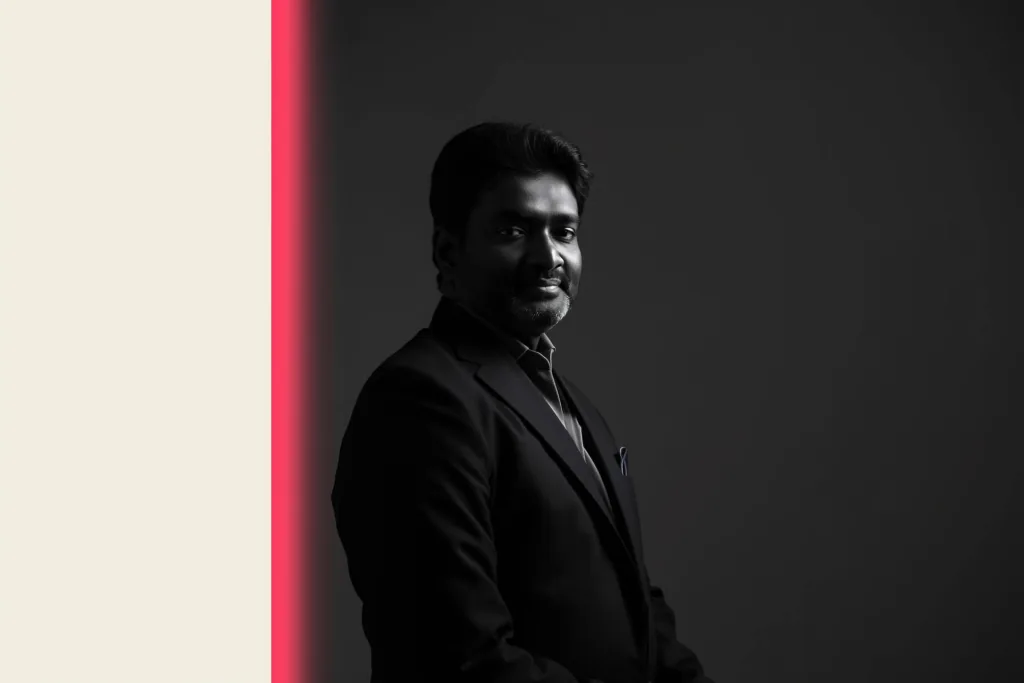 A confident Indian founder lit by a coral edge light in a dark office.