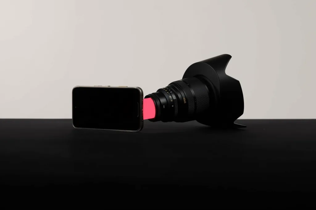 A smartphone and a cinema lens bridged by coral light on a dark surface.